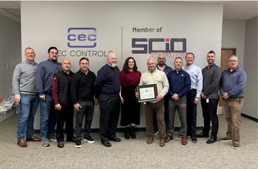 A group of eleven people stand in a row inside an office lobby in front of a wall featuring the logos for CEC Controls and SCIO Automation. One person near the center holds a framed award, while the others stand beside them, dressed in business‑casual attire.