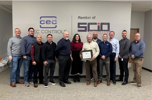 A group of eleven people stand in a row inside an office lobby in front of a wall featuring the logos for CEC Controls and SCIO Automation. One person near the center holds a framed award, while the others stand beside them, dressed in business‑casual attire.